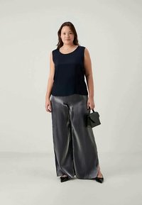 Γυναίκα με αμάνικο μαύρο τοπ, φαρδιά γυαλιστερά γκρίζα παντελόνια, μαύρα μυτερά παπούτσια και κρατώντας μια μικρή μαύρη τσάντα, ενώ στέκεται μπροστά σε απλό φόντο.