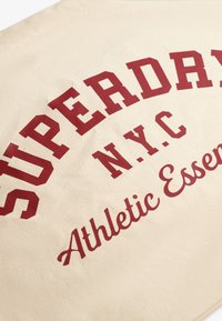 Sac en toile beige avec un texte audacieux et rouge foncé : "SUPERDRY N.Y.C." et "Essentiels Athlétiques". Le texte a un style vintage.