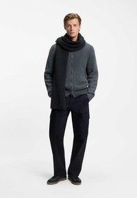 Grå ribbet cardigan med et mørkegrå tørklæde, parret med sorte cargo-bukser og sorte sneakers. Strikstrukturen er synlig på cardiganen og tørklædet.