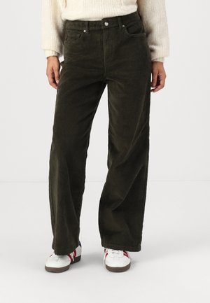 Person trägt dunkelgrüne Cord-Hose mit weitem Bein, weiße Turnschuhe mit roten Streifen und einen cremefarbenen Strickpullover.