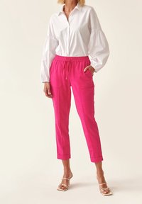 Jasně růžové tepláky s nastavitelným pasem a dvěma předními kapsami, v kombinaci s bílou košilí s knoflíky, která má nabrané rukávy.