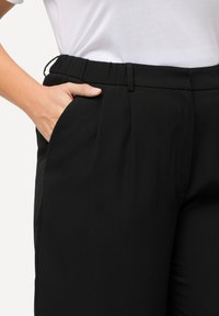 Zwarte broek met een brede elastische tailleband, een voorkeurszak en subtiele plooien. De stof lijkt glad en lichtgewicht te zijn.