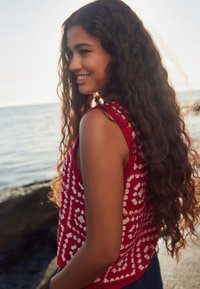 Rode gehaakte tanktop met witte bloemmotieven, voorzien van een mouwloos ontwerp en een ontspannen pasvorm. Op de achtergrond is een strand en water te zien.