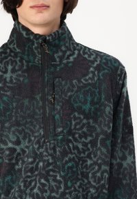 Mężczyzna noszący polarową kurtkę w ciemnozielono-czarne wzory z zamkiem błyskawicznym przy wysokim kołnierzu oraz kieszonką na zamek błyskawiczny na piersi.