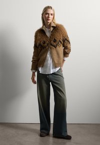 Brauner Strick-Cardigan mit Kunstfellverzierungen, schwarzen Akzenten und Reißverschluss. Kombiniert mit hellen Denim-Hose im weiten Schnitt und einem weißen Hemd.