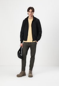 Black bomber jacket, light yellow t-shirt, olive green pants, beige lace-up boots, holding a black bag with white branding. Plain background.