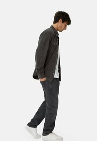 Mørkegrå skjorte med knapper og to brystlommer, båret over en hvid t-shirt, parret med mørke jeans og hvide sneakers, set fra en vinkelprofil.