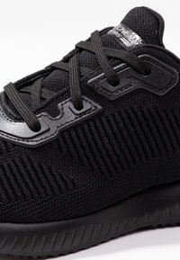Svart atletisk sko med ett texturerat tygöverdel, med förstärkta snörhål, svarta snören och en dämpad sula med ribbade detaljer.