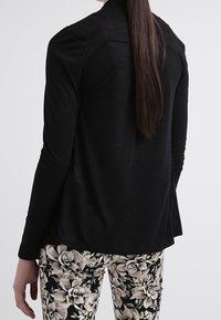 Schwarzes Langarmoberteil mit lockerer Passform und glatter Textur, kombiniert mit einer floral gemusterten Hose in Creme- und Grüntönen.