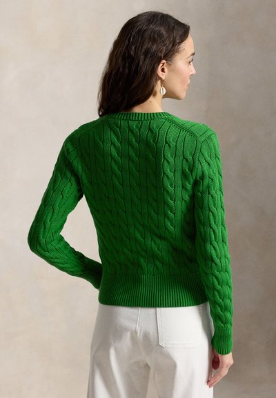 Žena s tmavými vlasmi má na sebe zelený sveter s vrkočovým vzorom a biele nohavice, stojí bokom s otočenou hlavou a chrbtom k neutrálnej stene.