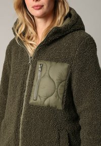 Olijfgroen fleecjack met een gestructureerde buitenkant, voorzien van een gewatteerde patchzak en een ritsluiting. Met capuchon voor extra warmte.