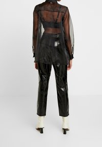 Černá průsvitná košile s dlouhým rukávem přes přiléhavé černé tričko, doplněná lesklými černými kalhotami a bílými kotníkovými botami s masivním podpatkem.