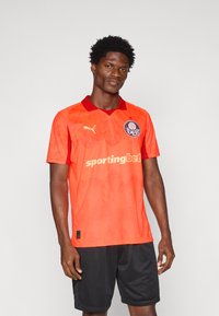 Svetlo oranžna nogometna majica z rdečimi poudarki in ovratnikom. Ponaša se s poudarjenim logotipom in teksturiranim vzorcem. Nosi se z črnimi hlačami.