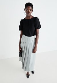 Top nero con maniche a sbuffo abbinato a una gonna grigia chiara plissettata con dettagli metallici. La modella tiene in mano una piccola borsa nera e indossa scarpe a punta.