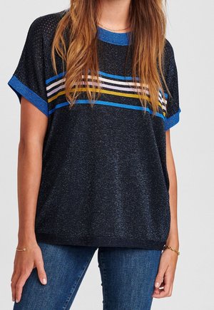 Žena v černém krátkém svetru s modrými, bílými a žlutými vodorovnými pruhy, v kombinaci s modrými džíny a zlatými náramky.