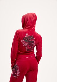 Piros velúr melegítőruha kapucnival. Mind a felsőn, mind az alsón nagyméretű, fekete csillogó felirat és díszítő minták találhatók a hátoldalon.