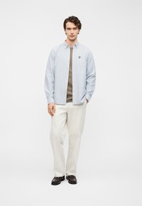 Ανοιχτό μπλε ριγέ πουκάμισο με κουμπιά πάνω από γκρι μπλουζάκι, συνδυασμένο με λευκό παντελόνι και σκούρα λοφιά· διαθέτει τσέπη στο στήθος.