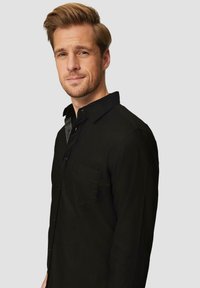 Schwarze Button-Down-Hemd mit Kragen, ausgestattet mit einer einzigen Brusttasche und einer glatten Textur. Getragen von einem männlichen Modell vor einem schlichten Hintergrund.