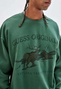 Groene sweatshirt van katoen met een ronde hals, voorzien van een afbeelding van twee herten en de tekst "GUESS ORIGINALS" en "VERKEN DE BUITENLANDSCHAP."