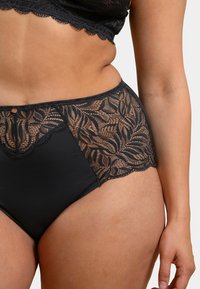 Culottes noires taille haute avec un overlay en dentelle florale sur les côtés et un tissu lisse. Présente des bords festonnés et un petit bouton décoratif.