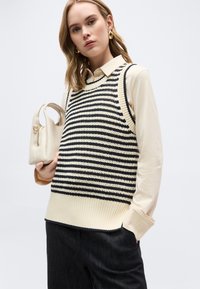 Νεαρή γυναίκα με πλεκτό γιλέκο σε ασπρόμαυρη ριγωτή σχεδίαση πάνω από κρεμ πουκάμισο, κρατώντας μικρή μπεζ τσάντα, με χρυσούς κρίκους σκουλαρίκια.