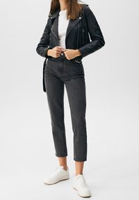 Veste de motard en cuir noir portée sur un t-shirt blanc, associée à un jean gris taille haute et des baskets blanches, posant devant un fond clair.