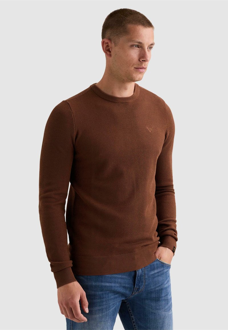 Homme aux cheveux courts portant un pull texturé marron et un jean bleu, debout avec une main dans la poche devant un fond uni.