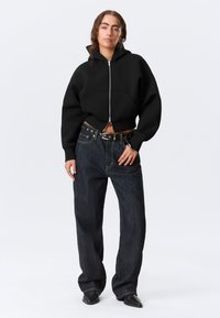 Μαύρο φούτερ cropped με φερμουάρ, χοντρό ύφασμα και μπροστινή τσέπη, σε συνδυασμό με σκούρα μπλε ευρύχωρα τζιν παντελόνια με ζώνη στη μέση.