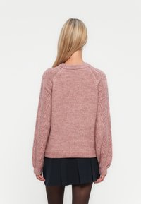 Donna con lunghi capelli biondi che indossa un maglione rosa lavorato a maglia con maniche decorate e una gonna nera plissettata, di spalle su sfondo bianco.