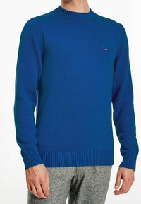 Mężczyzna ubrany w dopasowany niebieski sweter dzianinowy z prążkowanymi mankietami i dołem, w połączeniu z szarymi teksturowanymi spodniami.