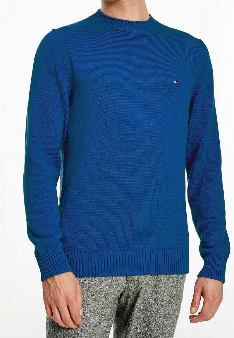 Mężczyzna ubrany w dopasowany niebieski sweter dzianinowy z prążkowanymi mankietami i dołem, w połączeniu z szarymi teksturowanymi spodniami.