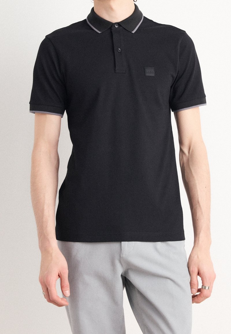 Homme portant un polo noir à manches courtes avec un liseré gris au col et aux manches, associé à un pantalon gris clair.