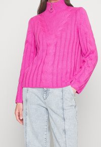 Pull tricoté fuchsia à col haut, avec des détails en maille torsadée, une texture côtelée et un tirette circulaire. Associé à un denim clair.
