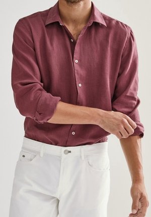 Chemise en lin bordeaux avec col, fermeture à boutons sur le devant et manches retroussées. Associée à un pantalon blanc, mettant en avant une coupe décontractée et une texture.
