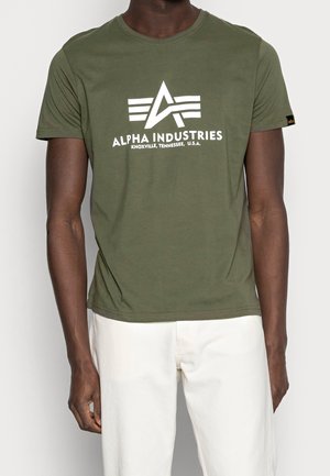 Olivengrøn bomulds-T-shirt med rund hals og korte ærmer, med et hvidt trykt logo og tekst foran. Hvide bukser.