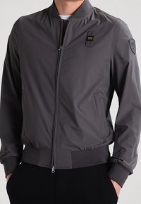 Gray bomber jacket made of smooth fabric, featuring a zip closure, ribbed cuffs, and a badge on the left shoulder.