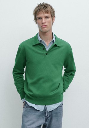 Giovane uomo con capelli arruffati che indossa una polo verde a maniche lunghe sopra una camicia azzurra, con le mani nelle tasche dei jeans grigi.