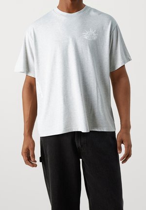 Ljusgrå kortärmad t-shirt med rund halsringning, prydd med en grafisk design på vänster bröst och med lös passform. Svarta jeans synliga längst ner.