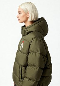 Veste matelassée vert olive avec capuche, arborant un graphique de main blanche et un détail de cœur rouge. Elle présente un design matelassé et des poches latérales.