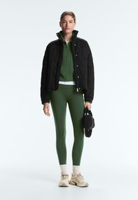 Kvinde står iført en sort quiltet jakke over en grøn lynlåstrøje og leggings, holder en sort taske, iført beige sneakers og hvide sokker.