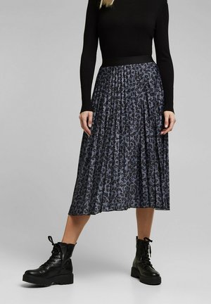 Mujer con camiseta negra de manga larga, falda midi plisada con estampado de leopardo gris y negro, y botines negros con cordones, de pie sobre un fondo gris.