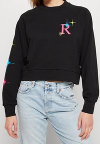 Sweatshirt corto nero con orlo a coste, maniche lunghe a raglan e grafica scintillante multicolore. Tessuto morbido con una vestibilità rilassata.