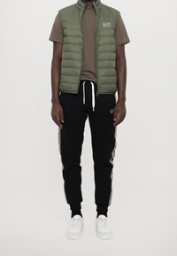 Zelená prošívaná vesta, hnědé tričko a černé tepláky s bočními pruhy. Bílá sneakers dotváří celkový outfit. Neformální a pohodlný styl.
