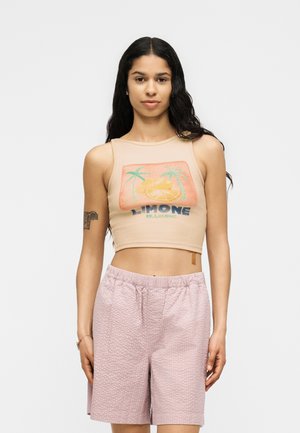 Ung kvinde med langt mørkt hår iført en beige crop top med palme- og citronmotiv og teksturerede lyserøde shorts.