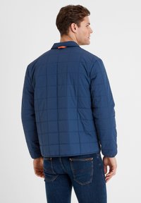 Navy quilted jacket with a structured design, featuring a collar and an orange accent at the back. Material appears smooth and lightweight.