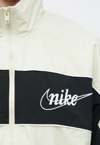 Μπουφάν Nike με ανοιχτό κρεμ χρώμα στο πάνω μέρος και μαύρο στο κάτω, με εμφανές λευκό λογότυπο και υφασμένη φερμουάρ.