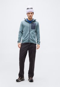 Νεαρός άνδρας που φοράει ένα ανοιχτό μπλε υφασμάτινο μπουφάν, μαύρο παντελόνι cargo, παπούτσια πεζοπορίας και λευκό σκούφο με μαύρο κείμενο, στέκεται μπροστά σε λευκό φόντο.