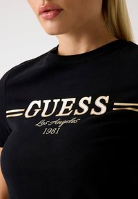 Čierne bavlnené tričko s vyšitým zlatým textom "GUESS Los Angeles 1981" a horizontálnymi akcentovými pruhmi na hrudi. Klasický dizajn s kruhovým výstrihom.