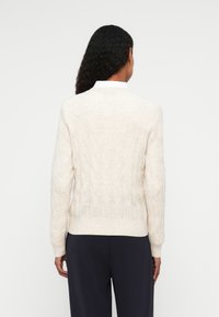 Crème kabelgebreide trui met lange mouwen en een geribbelde zoom, voorzien van een gestructureerd patroon en een ronde halslijn, gecombineerd met donkere broek.