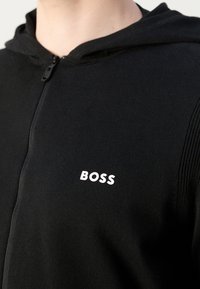 Schwarze Zip-Hoodie mit strukturiertem Design und einem weißen "BOSS"-Logo auf der Brust. Das Material ist weich und hat eine lockere Passform.